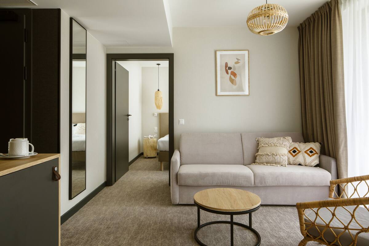 Ein Hotelzimmer mit einer Couch, einem runden Tisch, einem Korbsessel, einem Spiegel, einer Pendelleuchte und einem gerahmten Kunstwerk an der Wand.