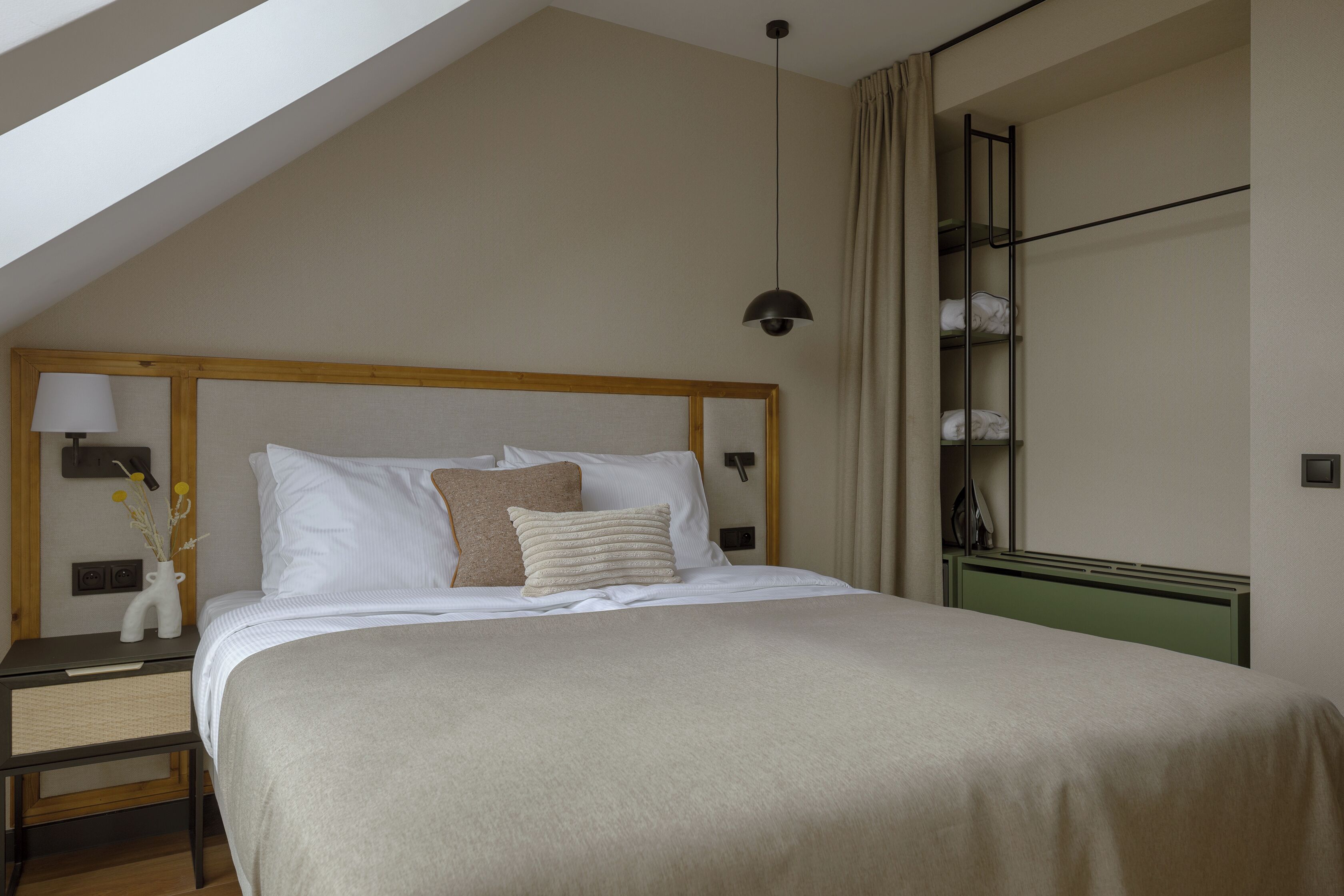 Une chambre avec un plafond en pente, un lit avec une literie blanche, une table de chevet avec une lampe et une étagère fixée au mur où sont pliées des serviettes.