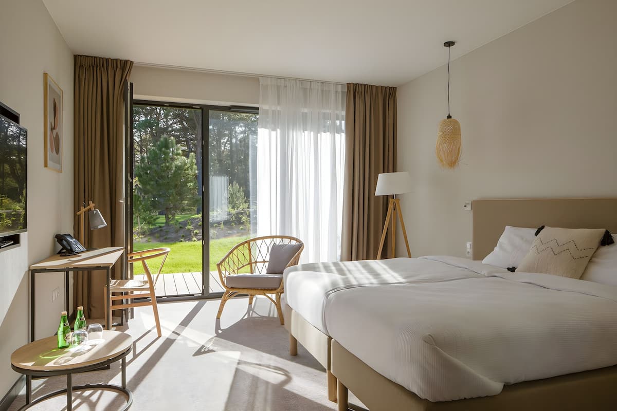 Ein Hotelzimmer mit Bett, Schreibtisch, Stuhl, einem kleinen Tisch mit Flaschen und Blick ins Freie.