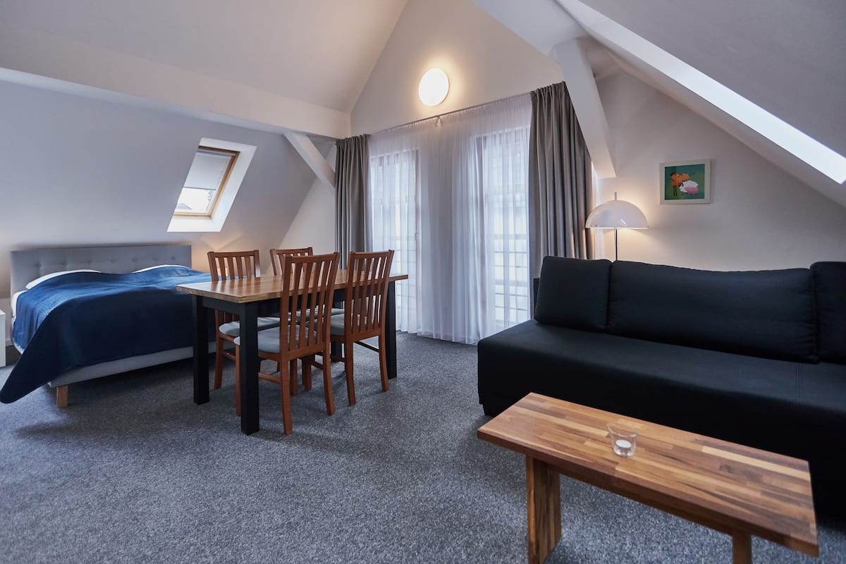 A room with a bed, a black sofa, a wooden table, a dining table with chairs, and a lamp.