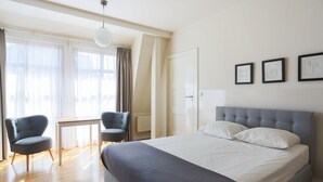 Ein modernes Hotelzimmer mit einem großen Bett, zwei Sesseln, einem kleinen Tisch und einem Fenster mit leichtem Vorhang.