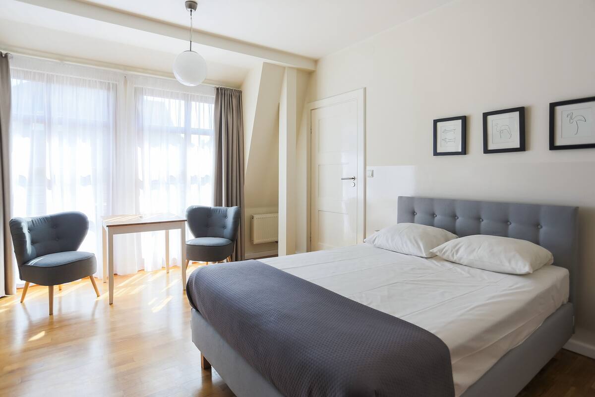 A modern hotel room with a large bed, two armchairs, a small table, and a window with sheer curtains.