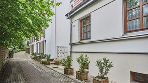 Ein schmaler, gepflasterter Weg, gesäumt von Gebäuden mit weißen Wänden und Holzläden. Entlang des Weges stehen mehrere Kübelpflanzen, und auf der rechten Seite ist ein kleines Fenster mit Blick auf Bäume zu sehen.