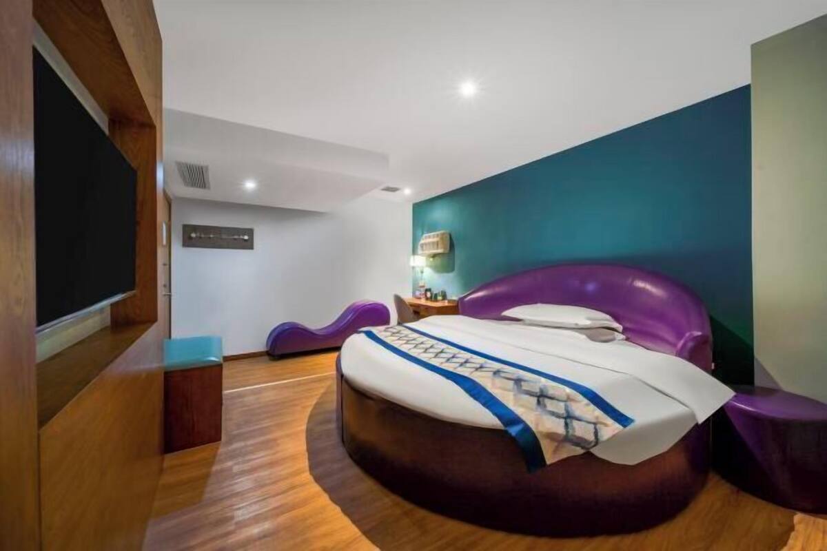 A modern hotel room with a curved bed, a flat-screen TV, a small desk, and a wall-mounted shelf.