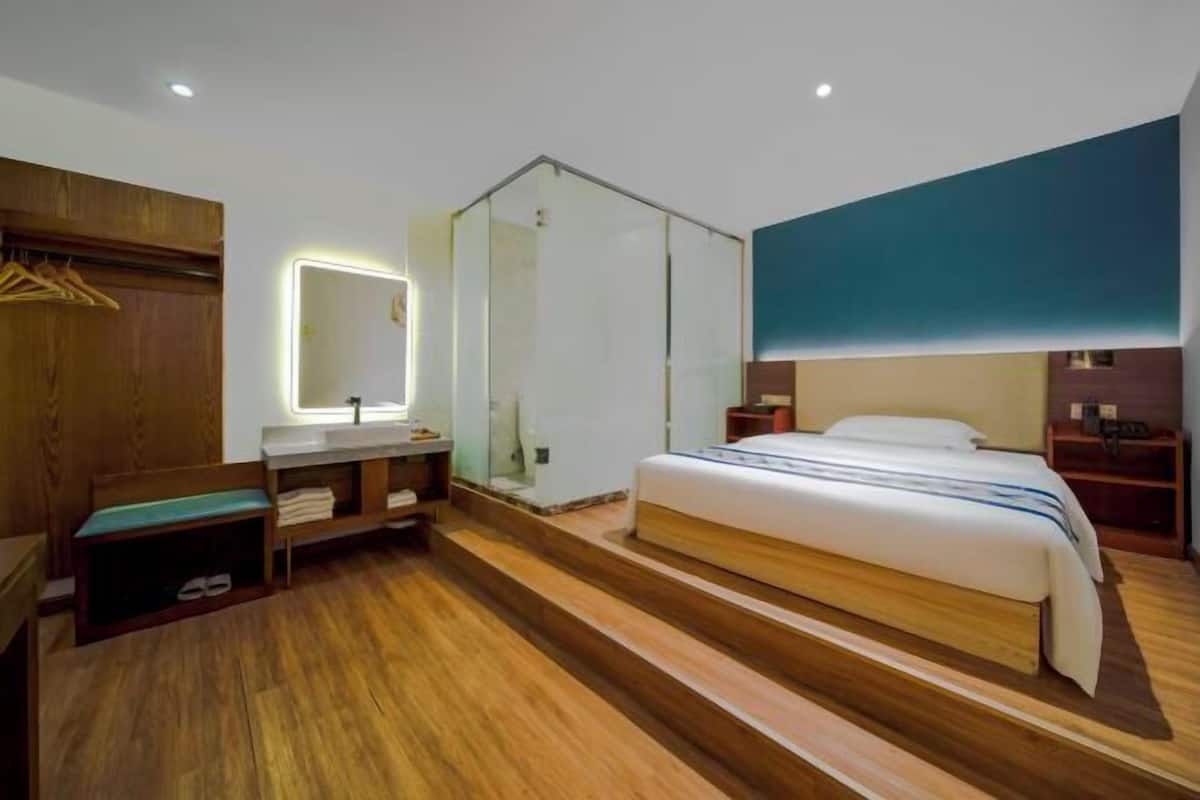 A modern hotel room with a wooden floor, a large bed, a glass shower enclosure, a bedside table, and a wall-mounted mirror.