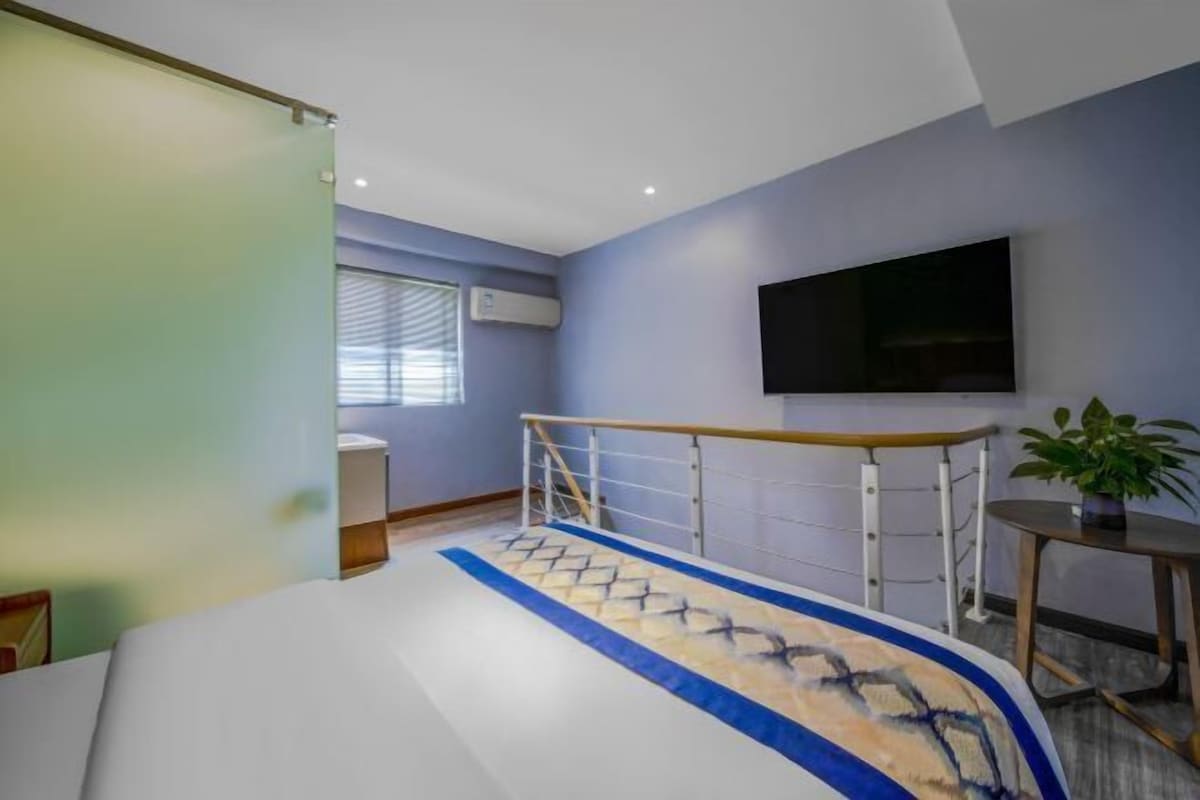 Een hotelkamer met een bed, een aan de muur bevestigde televisie, een raam met zonwering en een klein tafeltje met een plant.