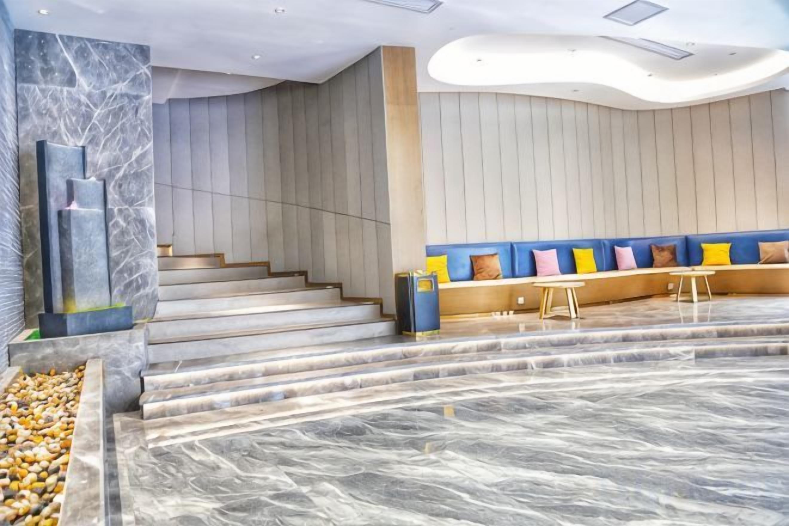 A modern hotel lobby with marble flooring, a staircase, a seating area with colorful cushions, and a reception desk.