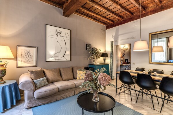 A living room with a sofa, a coffee table with flowers, a dining table with chairs, and artwork on the walls.