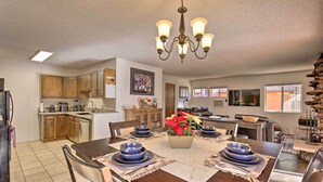 A dining area with a wooden table set for four, surrounded by chairs. A chandelier hangs above the table, and a potted plant with red flowers is placed in the center. - Hot Tub Hideaway in Pinetop: Bring Your Pet! (Pinetop-Lakeside)