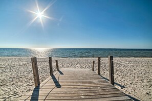 Une promenade en bois mène à une plage de sable fin, sous un ciel bleu azur et un soleil éclatant.