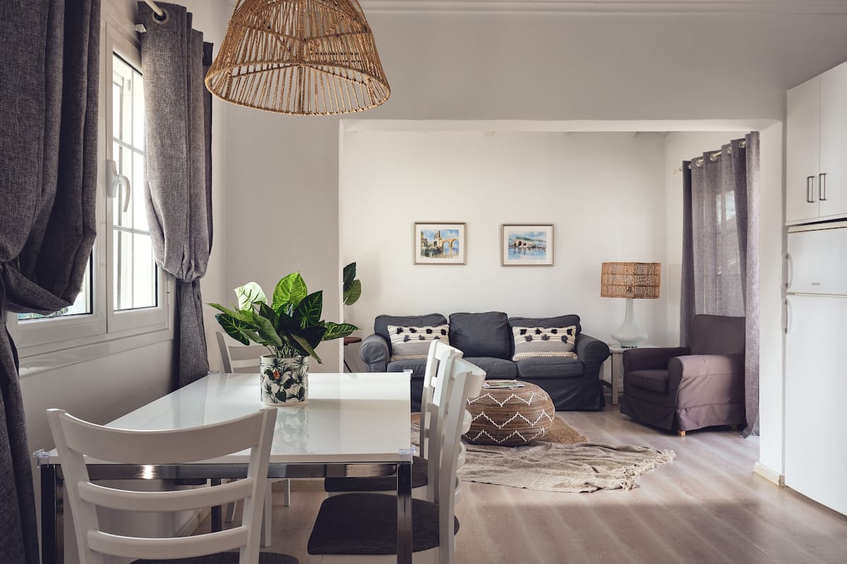 A modern living room with a dining area, featuring a white table, grey sofa, and a hanging light fixture.