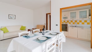 Ein modernes Wohnzimmer mit Esstisch, gedeckt für vier Personen, einer Couch und einer durchgehenden Tür sichtbaren Küche.