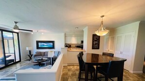 Ein modernes Wohnzimmer mit Sofa, Couchtisch und Fernseher. Es gibt einen Essbereich mit Tisch und Stühlen. Das Zimmer verfügt über ein großes Fenster und einen Deckenventilator.