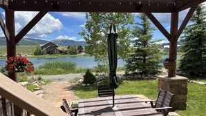 A wooden deck with a table and chairs, overlooking a lake with houses and mountains in the background. - Larkspur Luxury - Crested Butte (Crested Butte)