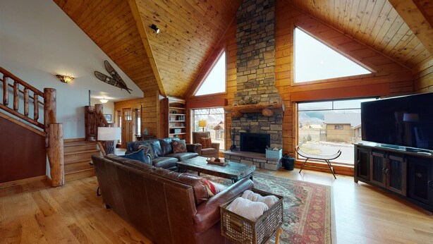 A spacious living room with a stone fireplace, wooden ceiling, and large windows offering a view of the outdoors. - Larkspur Luxury - Crested Butte (Crested Butte)