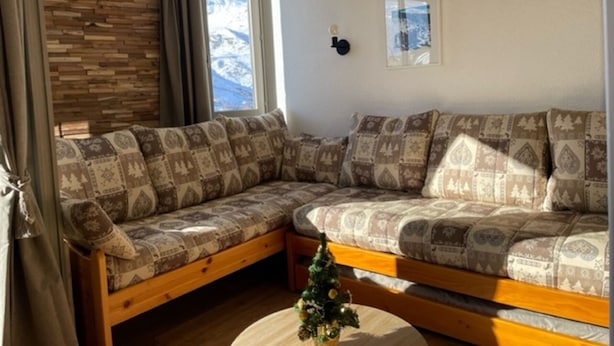 Un salon chaleureux avec un banc en bois et un canapé à motifs, un petit sapin de Noël et une fenêtre donnant sur des montagnes enneigées.