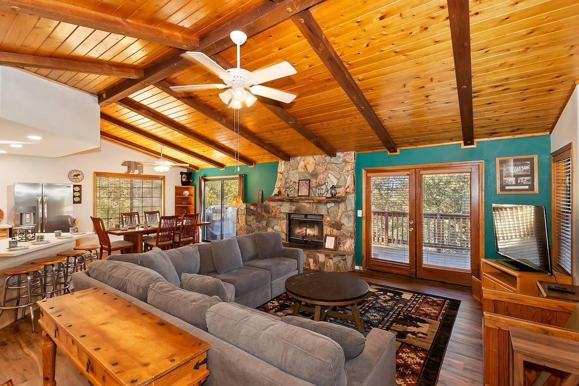 A spacious living room with a stone fireplace, wooden ceiling beams, and a large window overlooking a deck.