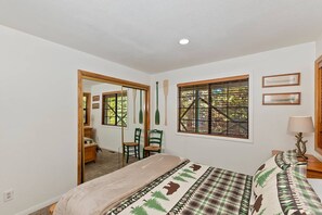 A bedroom with a bed, a window with blinds, a mirror, and a door leading to another room.