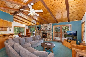 A cozy living room with a stone fireplace, a large gray sectional sofa, a wooden ceiling, and a television on a wooden stand.