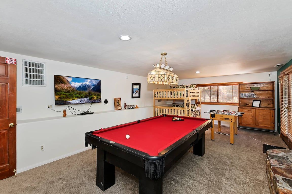 A pool table with a red felt surface, a television mounted on the wall, and a chandelier.