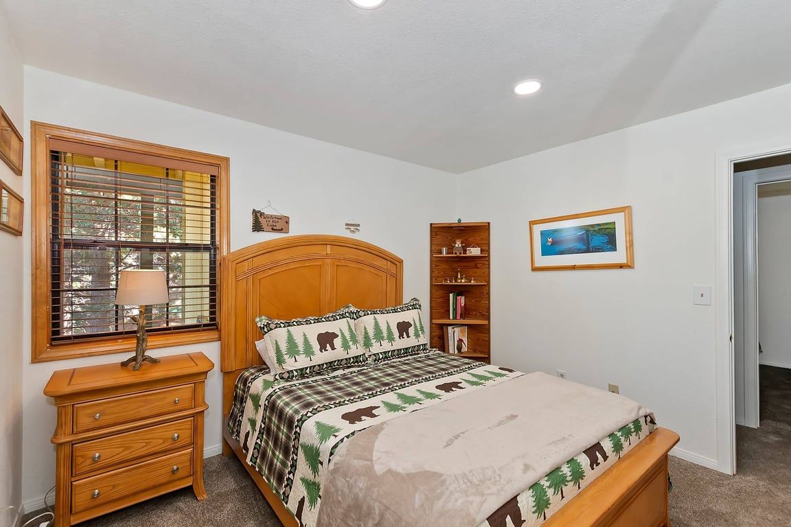 A bedroom with a bed, a nightstand, a lamp, a window with blinds, a bookshelf, and a framed picture on the wall.