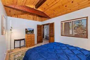 A bedroom with a wooden ceiling, a bed with a blue cover, a dresser, a television, and a window with blinds. - Castle Rock Cabin by Destination Big Bear : Located Close to the Lake! Hot Tub! Pool Table! Foosball! Fenced Yard! (Big Bear Lake)