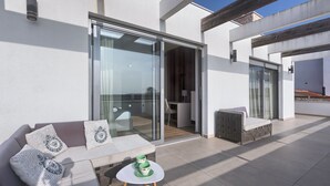 Ein moderner Außenbereich mit Sitzgelegenheit, einer Couch und einem Tisch, eine Glastür, die zu einem Innenraum führt, und ein klarer, blauer Himmel.