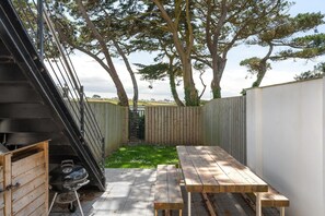 A patio with a wooden table and a barbecue grill, surrounded by a wooden fence and trees.