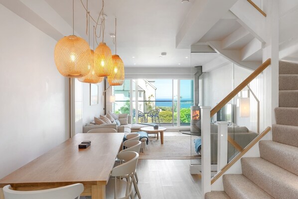 A modern interior with a dining area, staircase, and large windows overlooking the sea.