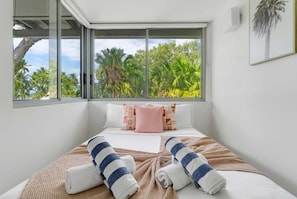 Une chambre d’hôtel avec un lit, une fenêtre donnant sur des palmiers et un tableau encadré au mur.
