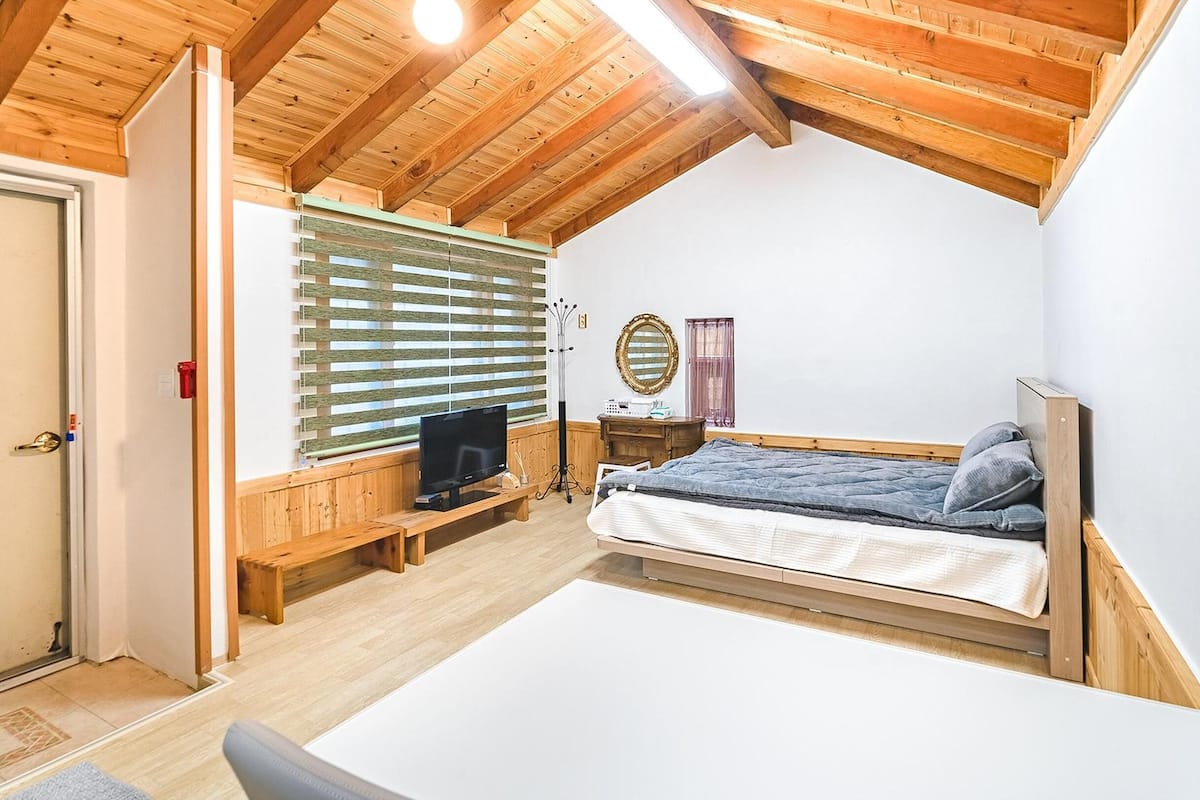 Ein Schlafzimmer mit Holzboden, einem Bett, einem Fernseher, einer Kommode und einem Fenster mit Jalousien.