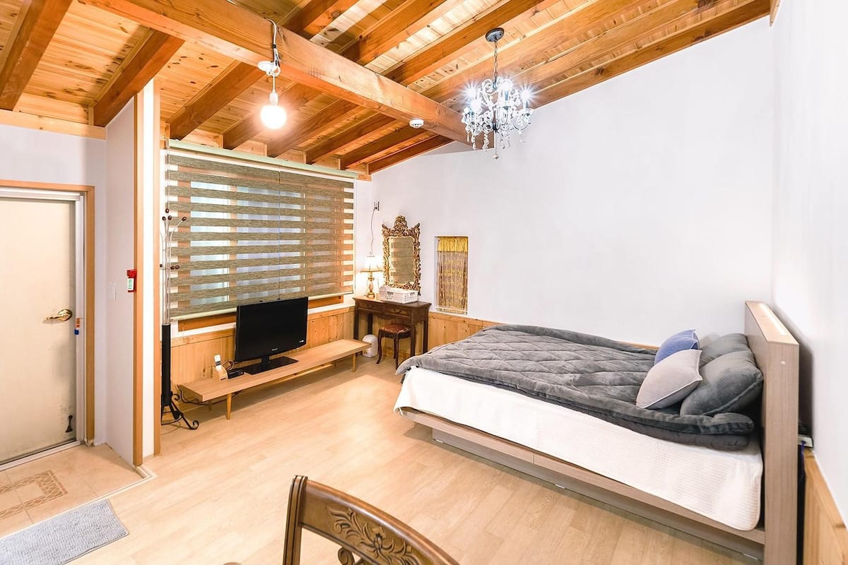 Ein Schlafzimmer mit einem Bett, Holzbalkendecke, einer Kristallleuchtre, einem Fernseher und einem Fenster mit Jalousien.