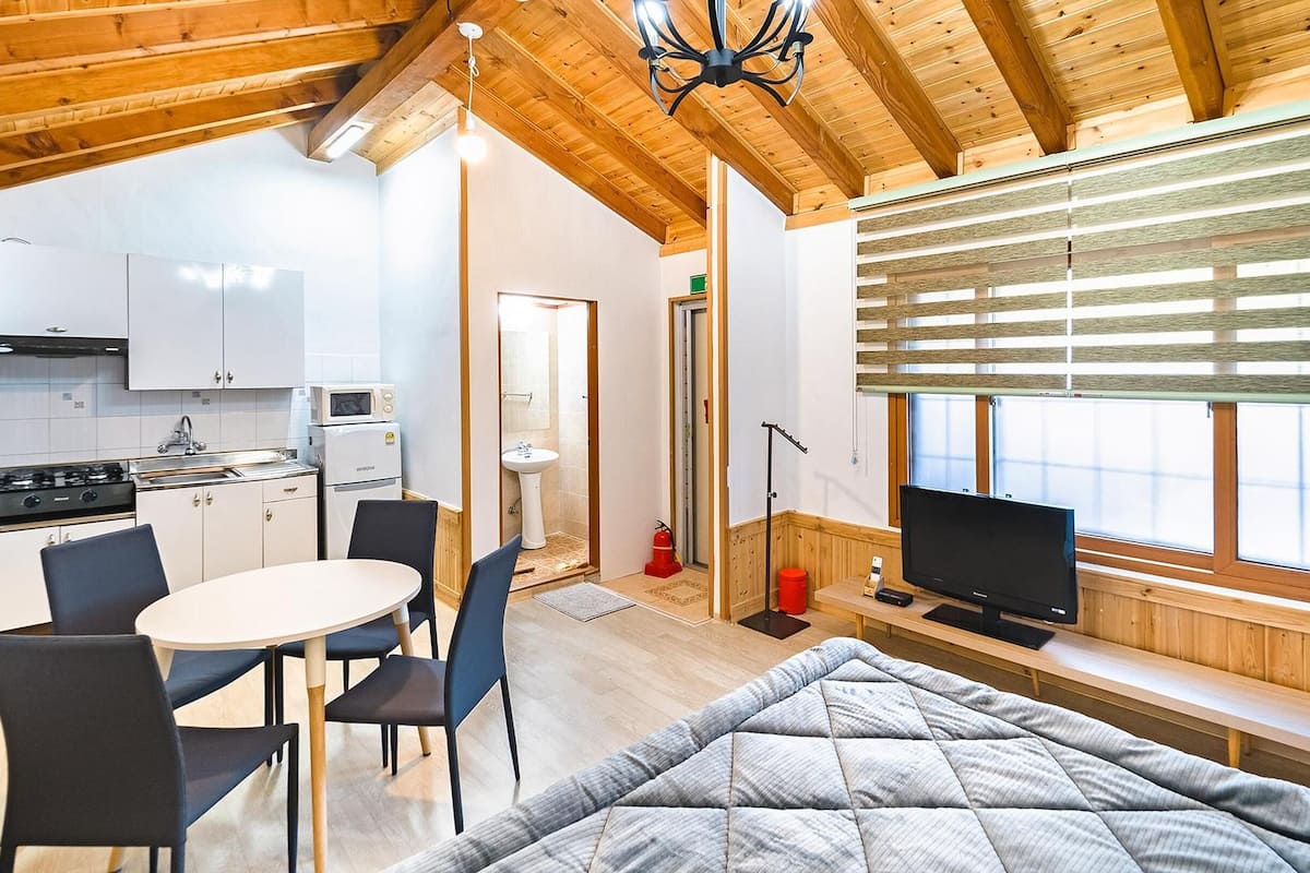 Ein gemütliches Zimmer mit einem Bett, einem Fernseher, einem Essbereich mit Stühlen, einer Küche mit Herd und Mikrowelle sowie einem Fenster mit Jalousien.