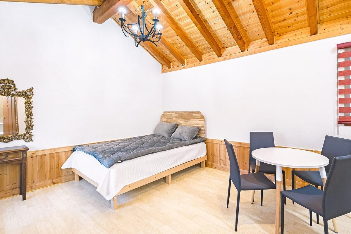 A room with a bed, a table with chairs, a chandelier, and wooden ceiling beams.