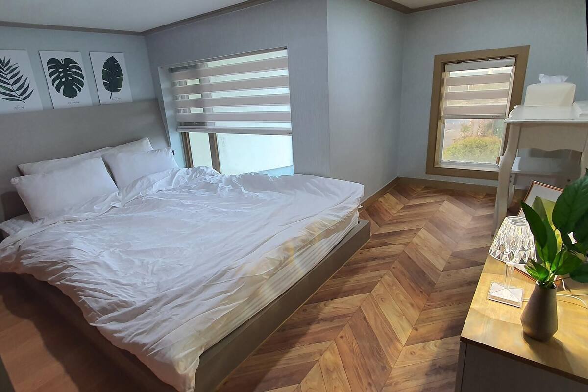 Une chambre à coucher avec un lit, un parquet en bois disposé en chevrons, une fenêtre avec des stores et une table de chevet agrémentée d’une plante.