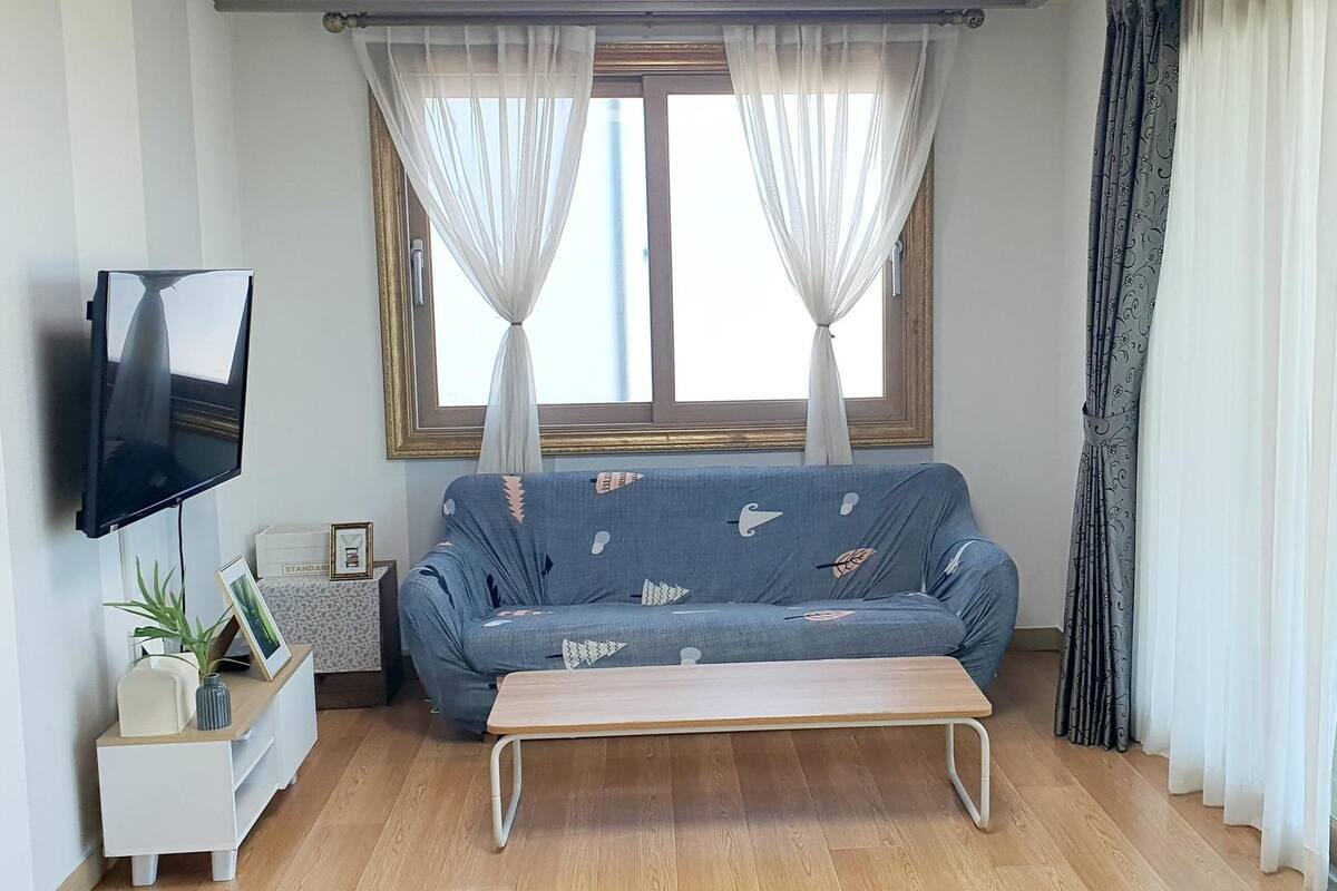 Un salon avec un canapé, un téléviseur, une petite table et une fenêtre avec des rideaux.
