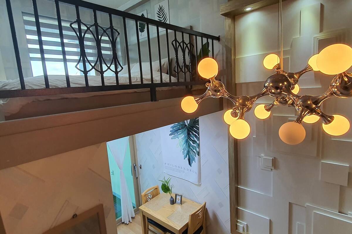 Une pièce comprenant un lit, une table à manger avec des chaises, un luminaire suspendu et un mur orné d’un motif de feuilles.