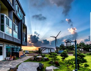 A modern building with large windows, a garden with rocks and trees, and a wind turbine in the background. - Jeju Basafan Spa Pension (Jeju City)