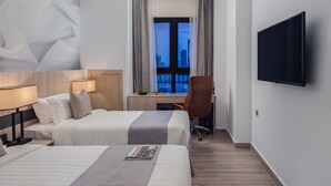 Habitación de hotel con dos camas, un escritorio, una silla, un televisor y un ventanal grande con cortinas.