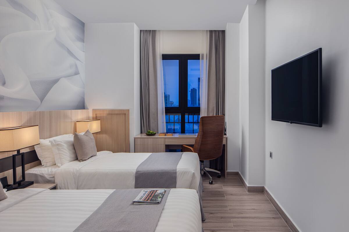 Camera d'albergo con due letti, una scrivania, una sedia, una televisione e un'ampia finestra con tende.
