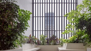 A modern indoor space with a view of a tall building through a grid of vertical blinds. There are multiple planters with various green plants and a built-in bench area.
