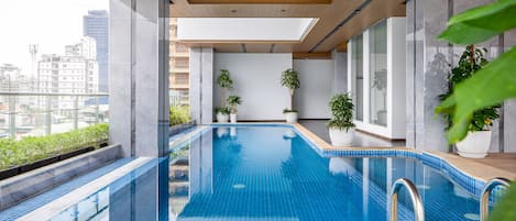 Piscina interior moderna con baldosas azules cristalinas, techo de madera y plantas en macetas.