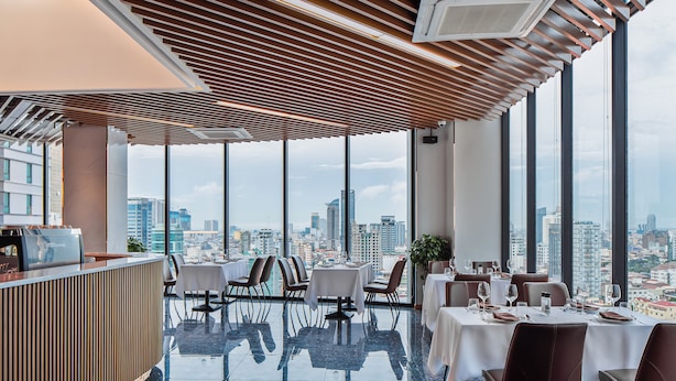 A modern restaurant with a city view, featuring a wooden ceiling, large windows, and a polished floor.