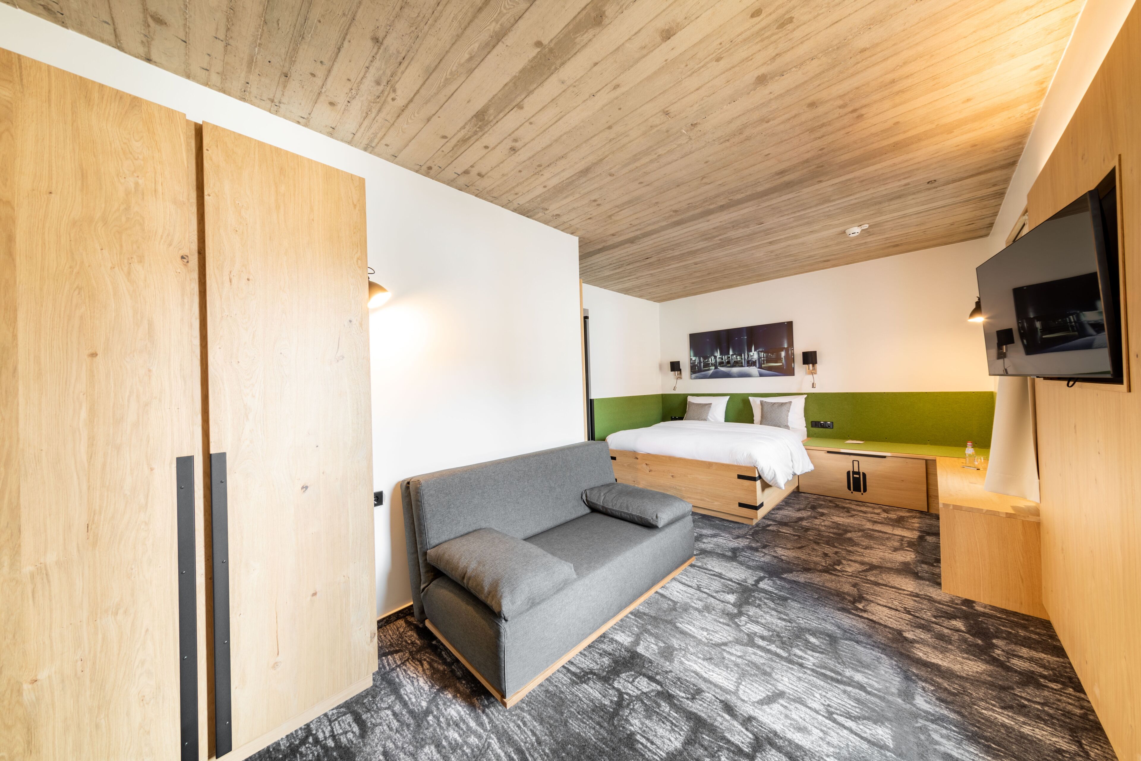 A modern bedroom with a wooden ceiling, a grey sofa, a bed with green headboard, and a mounted TV.