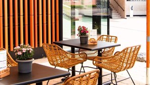 An outdoor seating area with wicker chairs and tables, a potted plant, and a view of a building through a glass wall. - De Brauhotel (Käerjeng)