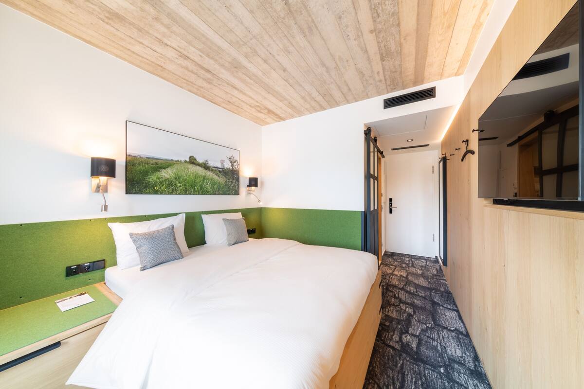 A modern hotel room with a large bed, a green accent wall, a framed landscape picture, and a wooden ceiling.