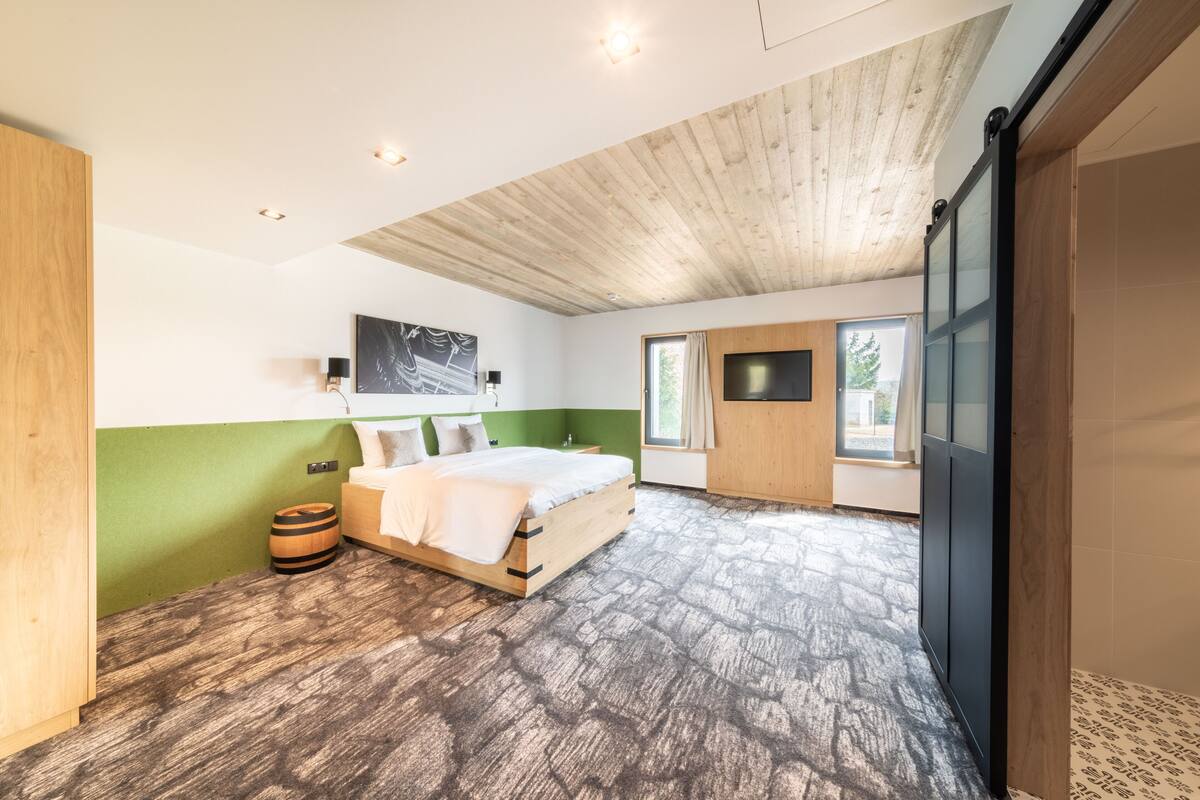 A bedroom with a wooden ceiling, a bed, a green wall, a window, and a TV.