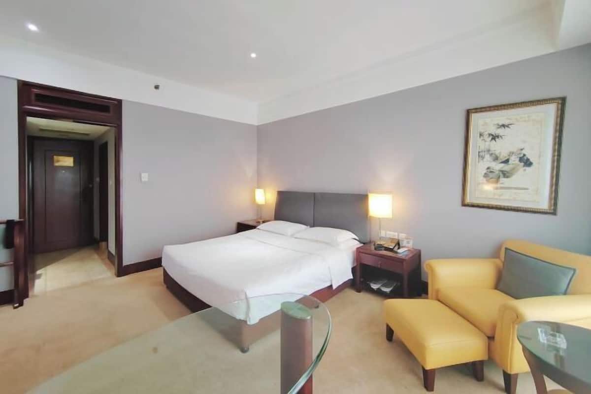 Une chambre d’hôtel avec un lit, des tables de chevet, une chaise, un pouf et un tableau encadré au mur.