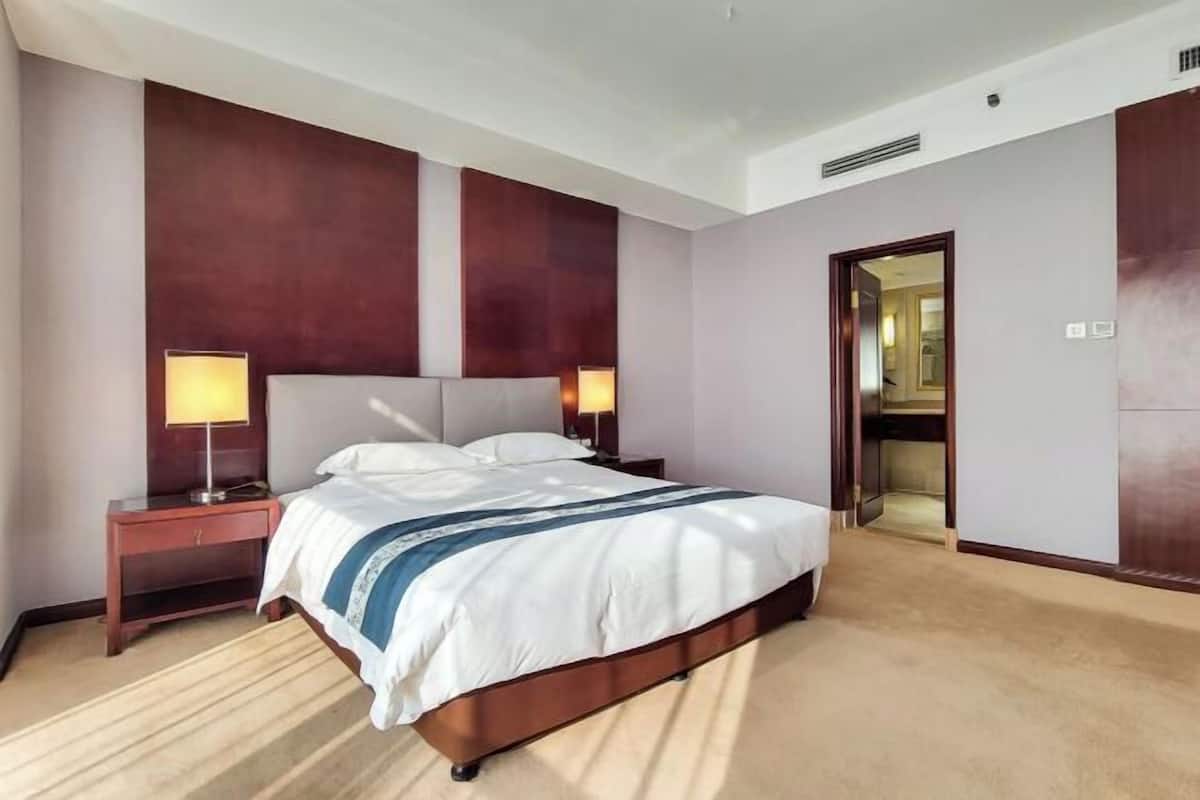 Une chambre d’hôtel avec un grand lit, deux lampes de chevet, une tête de lit en bois, un sol recouvert de moquette et une porte donnant sur une autre pièce.