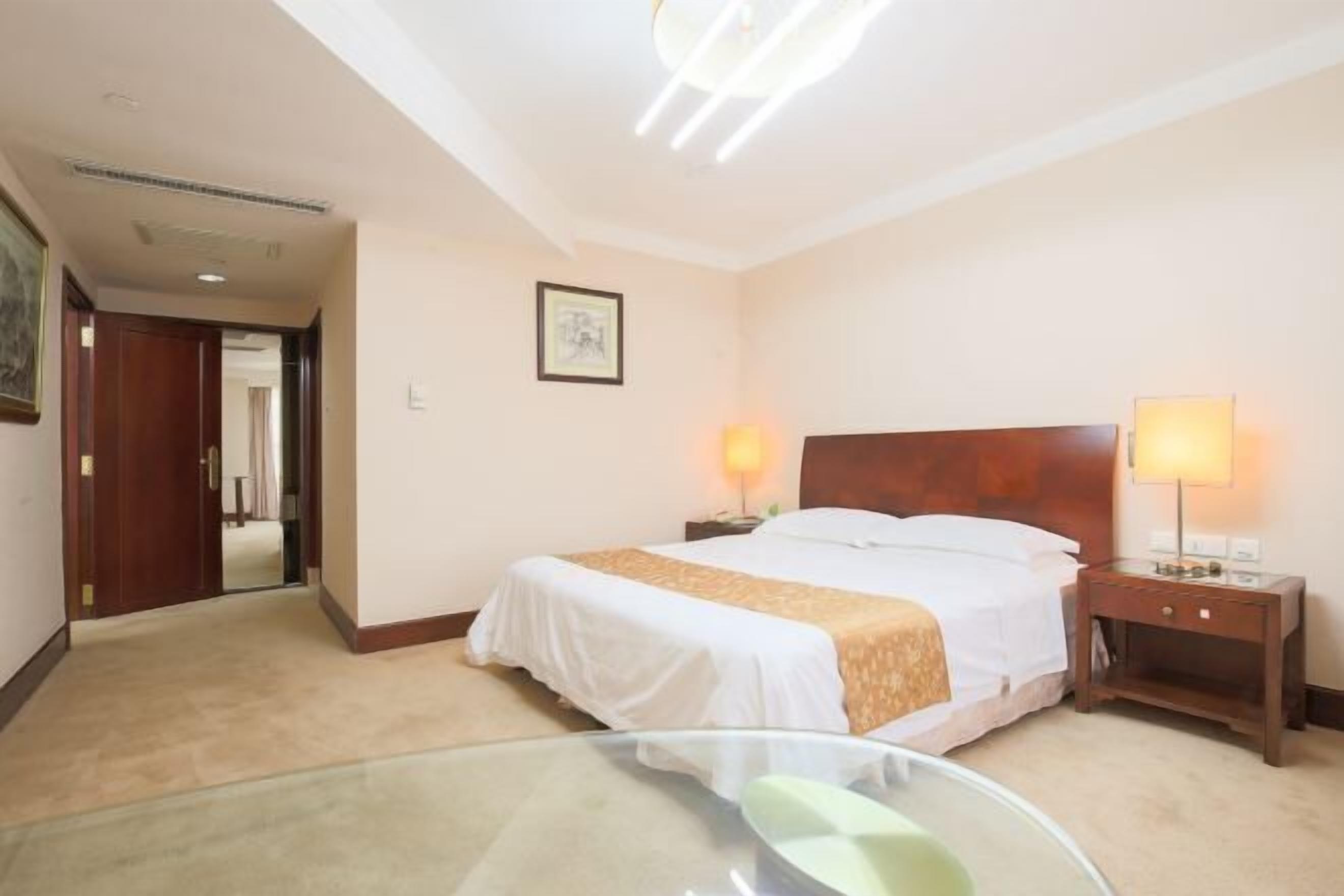 A hotel room with a large bed, two bedside lamps, a wooden headboard, a framed picture on the wall, and a glass table with a chair.